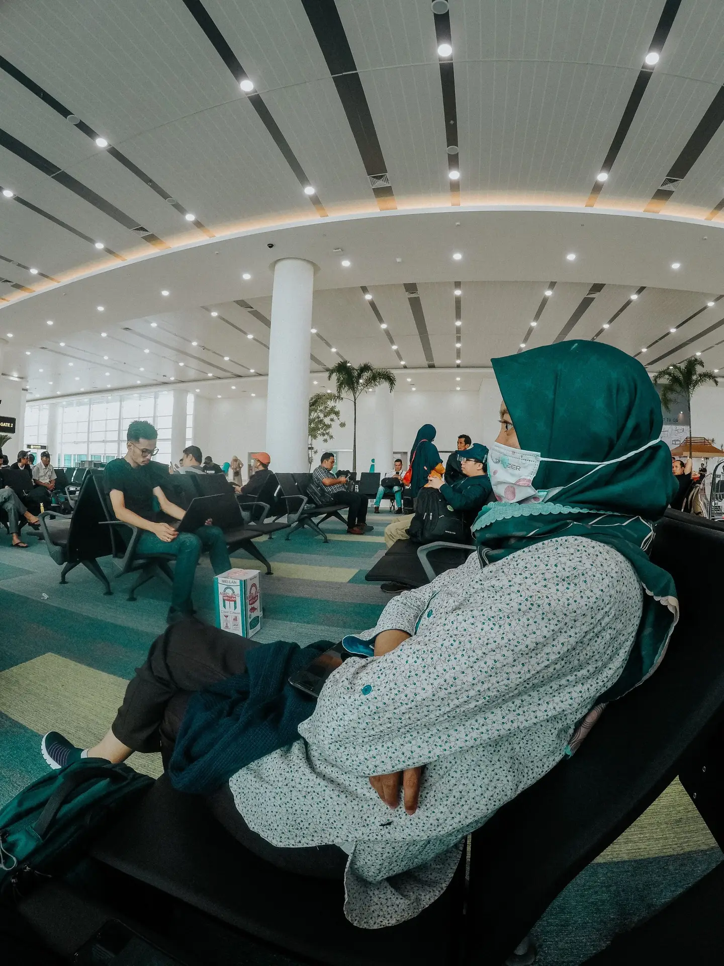 A woman in a hijab and mask patiently waits in a busy airport lounge, showcasing travel and safety.